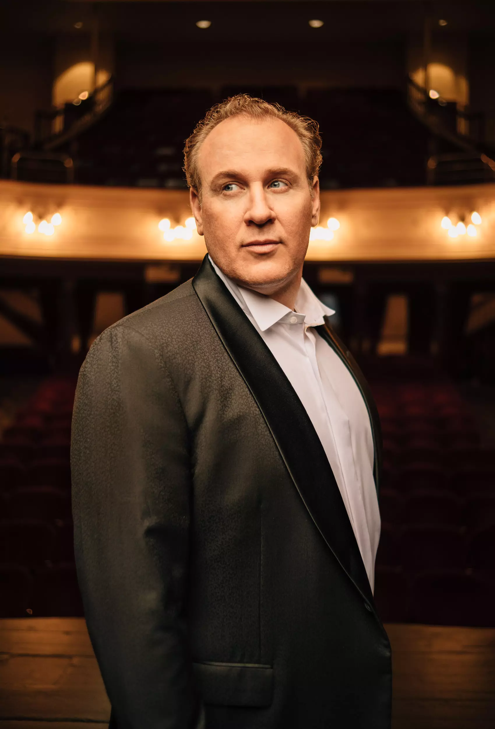Lucas Meachem, Baritone, dramatically-lit and wearing a tuxedo in an empty theatre, looking off to the left.